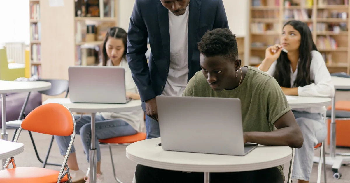 Estudiantes trabajando con laptops en una biblioteca mientras un tutor ayuda a uno de ellos