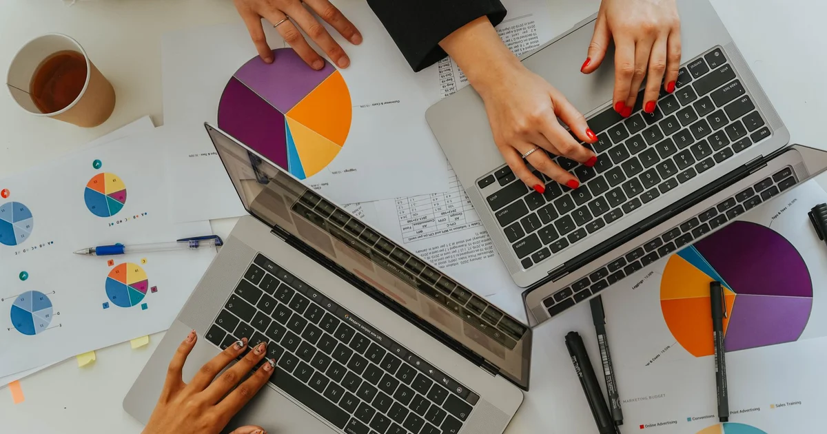 Vista cenital de una mesa de trabajo con dos laptops, manos escribiendo en los teclados e informes impresos con gráficos circulares de colores, bolígrafos y un vaso de café