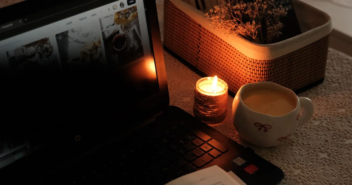 Laptop encendida junto a una vela y una taza sobre una mesa en penumbra