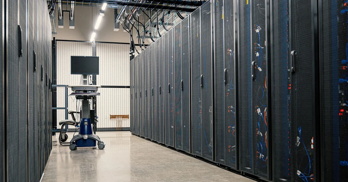 Pasillo de un centro de datos con filas de racks negros de servidores con puertas de malla y cables azules y naranjas, y al fondo un carrito con monitor