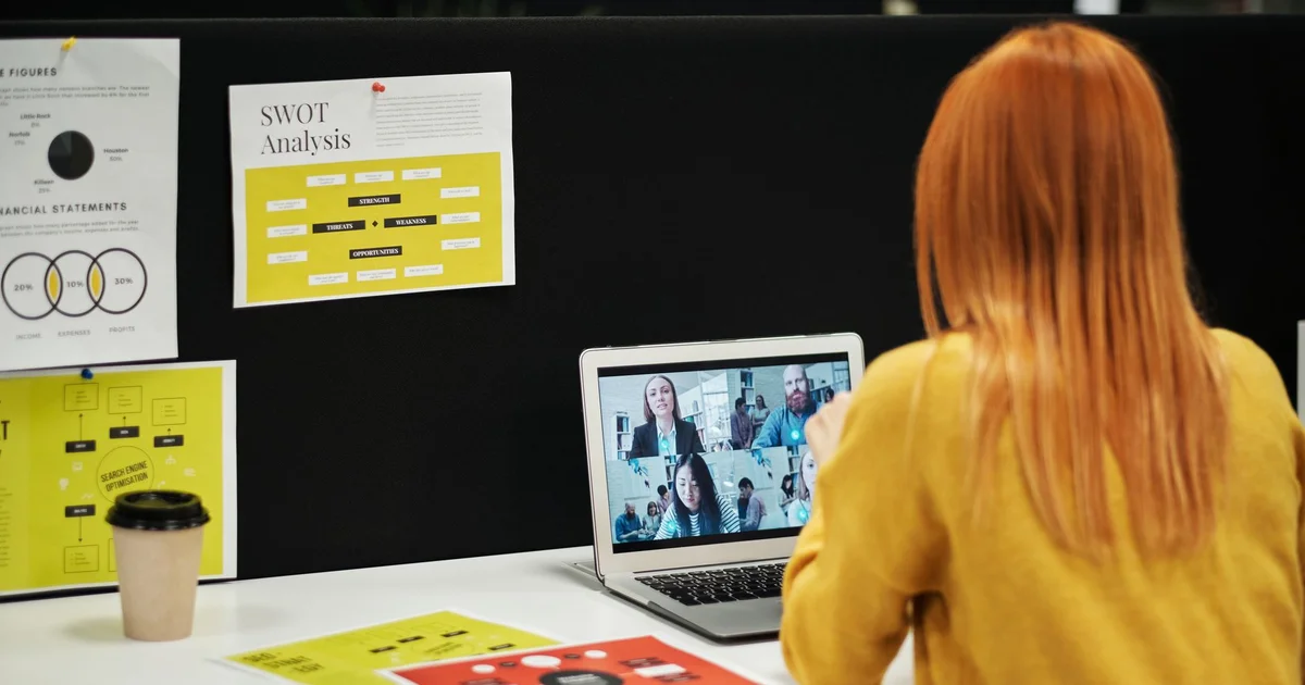 Mujer en una videollamada de equipo desde su laptop, frente a un tablero con gráficos de trabajo