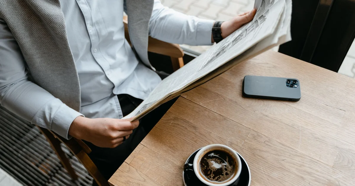 Hombre lee el periódico en la mesa de un café, junto a una taza de café y su celular